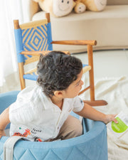 Child sitting in a blue chair holding a bottle in a room with a wooden chair