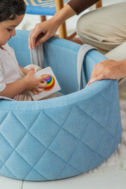 Child sitting in a blue baby bouncer with a toy, supervised by an adult.