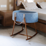 Blue quilted bassinet on a wooden stand in a bedroom setting